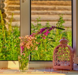 A bouquet of wildflowers in a glass and a beautiful pink candle holder on a windowsill in a farmhouse.