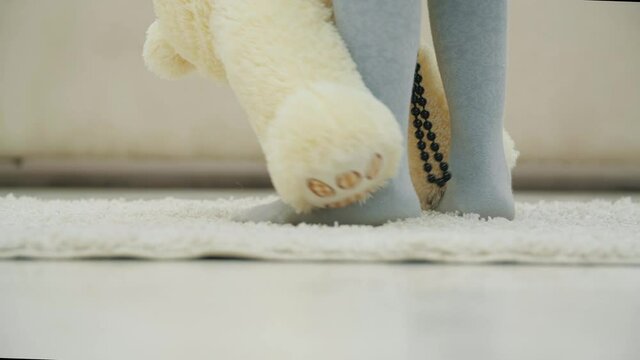Cropped legs of little girl standing on white carpet, holding a plush teddy bear in necklace, putting its paw on her feet and teaching it how to dance.