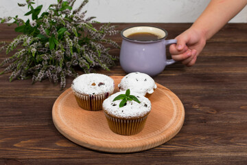 A child's hand holds a cup of tea and mint near the board with muffins with black currant, on a background of flowers and mint leaves on a wooden background