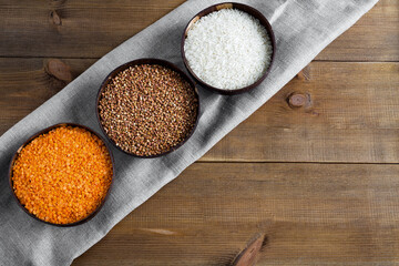 Three full bowls of raw cereals buckwheat, rice, lentils on a canvas napkin on a wooden background flat lay with place for text. Different types groats.