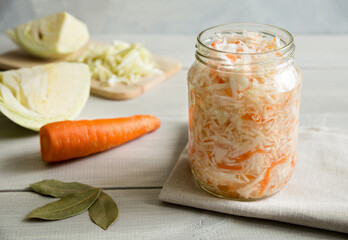 Open glass jar of sauerkraut stands on a napkin. Behind is a sliced fresh cabbage and carrots. Next to the bay leaf. Close up composition on a white, wooden background. Homemade salad. Fermented food