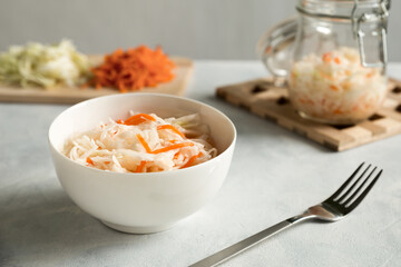 White ceramic bowl with homemade appetizer sauerkraut on a gray, concrete background close up. Glass jar and ingridients chopped coleslaw and carrot.