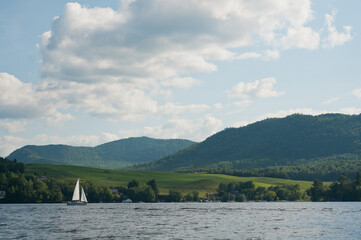 Horizontal landscape view of lush green mountains and fields and white sailboat on a partially cloudy summer day taken from lake Memphremagog.