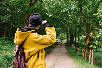 Back view of faceless female traveler with backpack walking along empty pathway surrounded by green trees on sunny day
