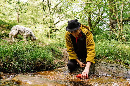 Stylish female in outerwear taking clean water from creek while resting in green forest with dog