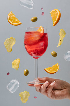 Crop female with glass of red alcohol cocktail garnished with orange slices and green olives above ground on gray background