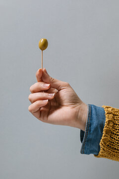 Unrecognizable crop female with green olive on toothpick for cocktail on gray background in studio