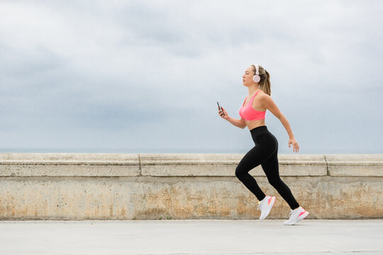 Side view of slim female runner in sports clothes and headphones jogging on pavement with cellphone under cloudy sky in daylight