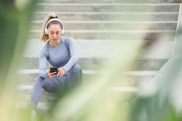 Thoughtful fit female athlete in headphones and sportswear resting on stairs after workout listening to music and browsing internet on cellphone in daylight
