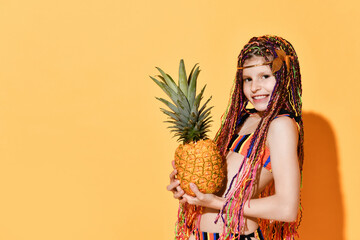 Lovely schoolgirl with cute african braids posing in striped bikini holding big pineapple close to cheek. Cropped shot isolated on orange, copy space