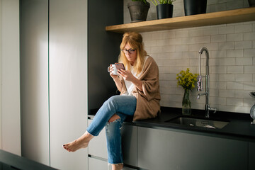 Serene female in cozy clothes sitting on counter in kitchen and reading messages on social media while enjoying fresh beverage in morning