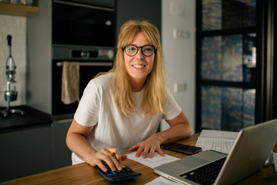 Happy female bookkeeper in casual outfit sitting at table in kitchen and counting bills while doing paperwork using calculator looking at camera