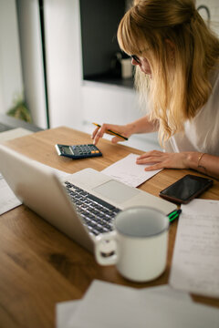 Focused female bookkeeper in casual outfit sitting at table in kitchen and counting bills while doing paperwork using calculator