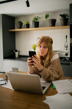 Side view of pensive female accountant sitting at table using on mobile phone while having break during remote work at home and laughing