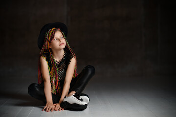 Fototapeta na wymiar Teenager with african pigtails, in black hat, leggings, mesh blouse and sneakers. Sitting on floor, posing on dark background