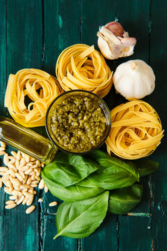 Top view of composition of dry pasta rolls near bowl with pesto sauce and fresh basil leaves with crushed garlic and pine nuts near bottle with olive oil