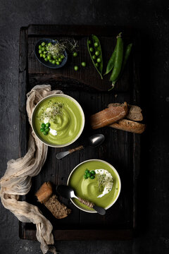 From above view of two bowls of pea cream placed on a wooden tray and dark surface, served with bread and decorated with whole peas and sour cream
