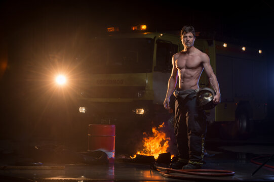 Muscular fireman with naked torso standing with helmet near flame and fire truck while looking at camera