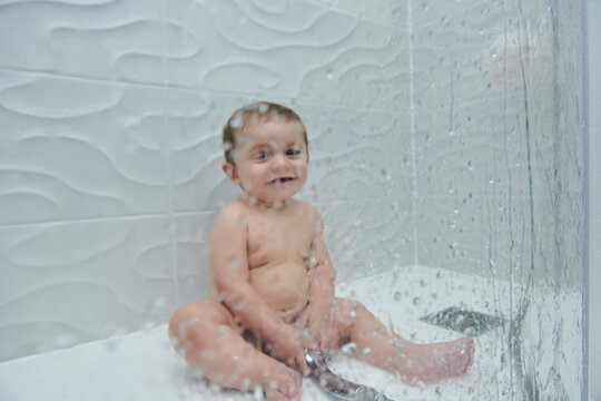 Cute cheerful little toddler sitting on floor of shower cabin and enjoying water procedure during bathing time at home