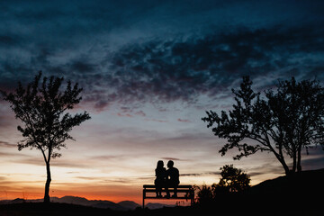 Back view silhouettes of affectionate couple enjoying romantic date while sitting on bench against cloudy sunset sky in summer evening