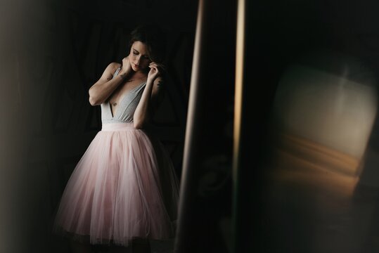 Calm graceful young female dancer in elegant gown touching hair gently while standing looking away in dark studio