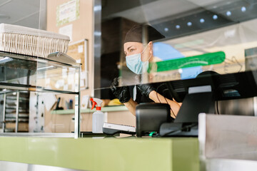 Positive cafe staff in uniforms and respirators working at cafe counter with cash register during coronavirus pandemic