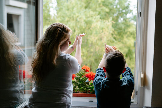 Back view of happy excited mother and son in casual clothes standing on opened window at home decorated with red potted flowers and clapping hands while looking out