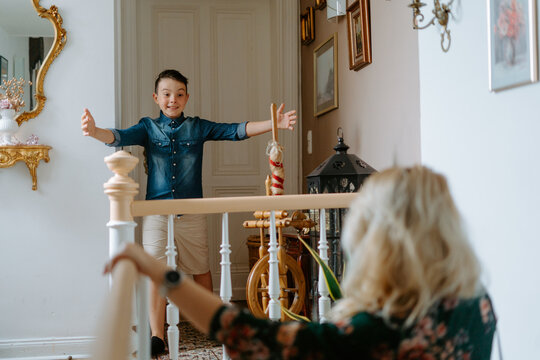 Happy excited grade schooler boy in casual wear standing in cozy hall near stairway with widely opened arms ready to embrace mother