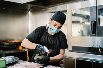 Concentrated female cook wearing latex gloves and medical mask spraying with oil chicken meat placed on roasting pan before baking