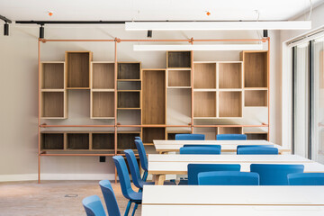Modern style office interior with square tables and blue stools on floor and wooden shelves on wall with large window and lamps on ceiling