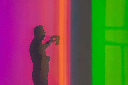 Shadow of anonymous male taking photo on smartphone while standing in multicolored neon studio