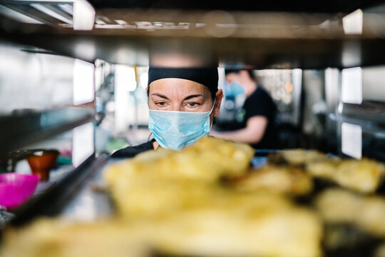 Concentrated female cook in hat and respirator standing against steel pan rack loaded with food while working in modern restaurant kitchen