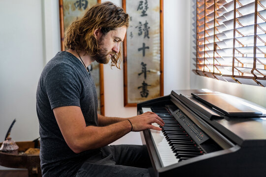 Side view of handsome male musician with long hair sitting in cozy apartment and playing electronic piano