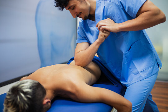 Smiling chiropractor with mustache in blue uniform putting elbow on back of anonymous patient lying on examination couch in clinic