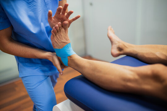 Top view of crop unrecognizable chiropractor in blue uniform attaching kinesio tape on foot of faceless man lying on examination couch in clinic