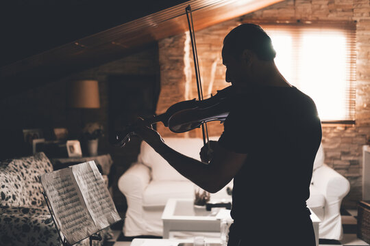Side view of silhouette of male musician playing violin and looking at music sheets while practicing in living room