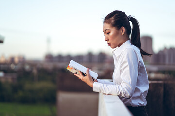 Side view of young Asian businesswoman in formal white shirt standing on rooftop of city building and reading book while relaxing after work