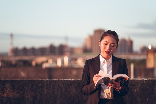 Front view of young Asian businesswoman in formal outfit standing on rooftop of city building and reading book while relaxing after work