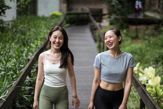 Smiling fit Asian girlfriends wearing tight casual clothes strolling along narrow wooden bridge in countryside and chatting