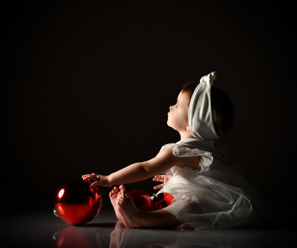 Baby Girl In White Headband And Dress, Barefoot. Holding Two Red Balls, Looking Up, Sitting On Floor. Twilight, Black Background.