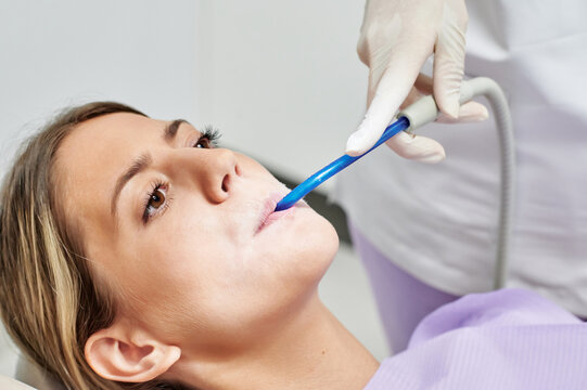 Faceless assistant of dentist holding salva ejector in mouth of female patient lying under lamp in hospital office