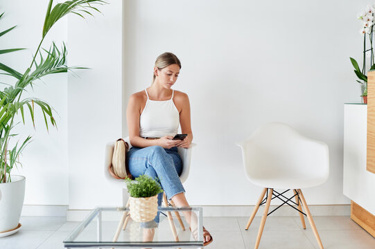 Full body of female client sitting on chair and browsing on smartphone while waiting for appointment in lobby of modern clinic