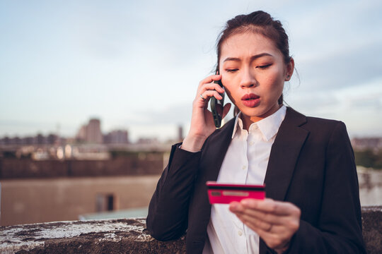 Serious Young Asian Businesswoman In Formal Wear Holding Credit Card And Having Phone Conversation With Bank Customer Service While Standing On Rooftop Of City Building