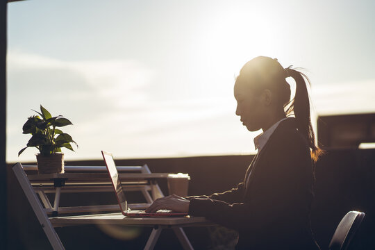 Side view of young Asian business lady in formal outfit using laptop while working remotely on rooftop terrace in city
