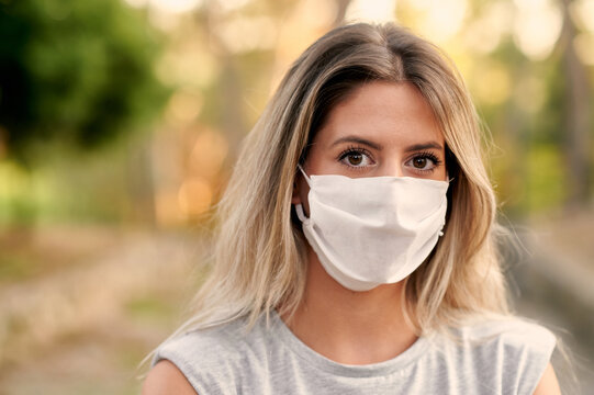 Young female in casual clothes and protective mask for coronavirus prevention looking at camera while standing on alley in green park