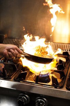 Cropped unrecognizable chef tossing frying shrimps in flambe above gas stove with burning flame while cooking in restaurant