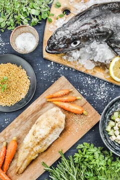 Top view of whole fresh raw fish on ice cubes and fried fillet with carrots on black table with spices and greens