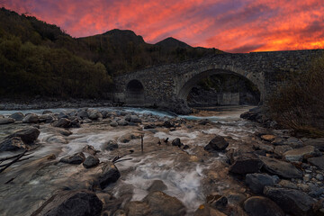 River and stony shore streaming through rocks and ancient stone bridge at sunset