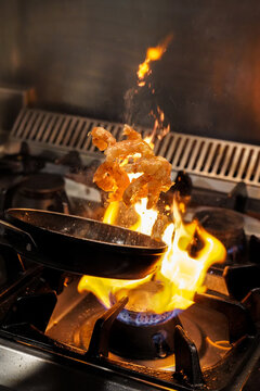 Chef tossing frying shrimps in flambe above gas stove with burning flame while cooking in restaurant