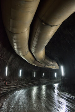 Wide passage in mining tunnel with glowing lamps on stone walls and huge metal pipe